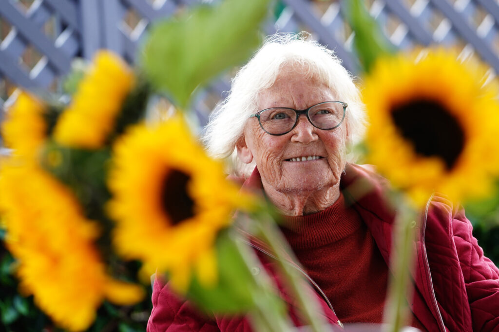 A woman (Margaret) smiles amongst sunflowers. 