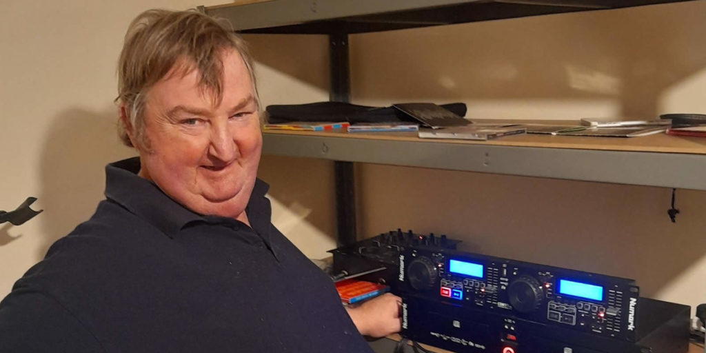 A man (Graeme) smiles towards the camera. There is DJ equipment in the background. 