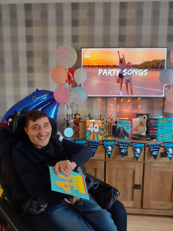 A man in a wheelchair smiles at the camera while holding a 40th Birthday card. The room is decorated in birthday balloons and bunting.