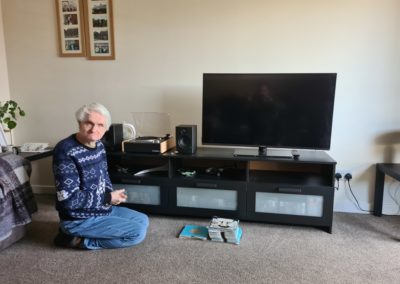 An older gentlemen kneels on the floor in front of a tv and record player.