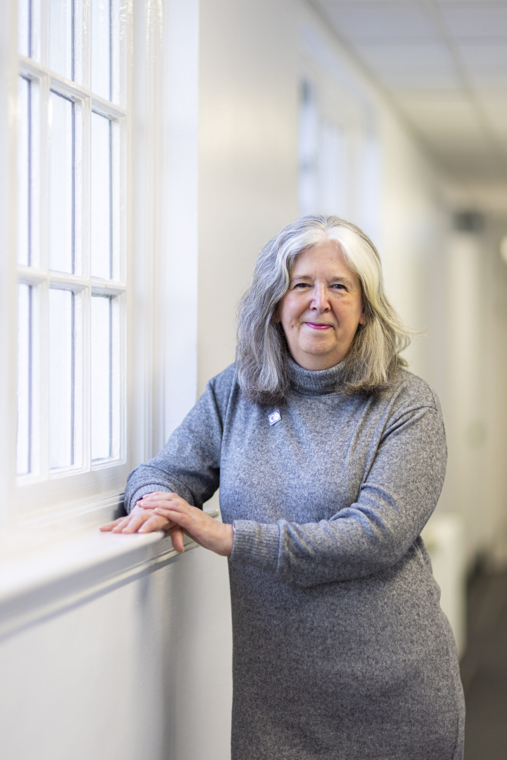 A woman (Lucy) standing by a window, wearing a gray sweater.