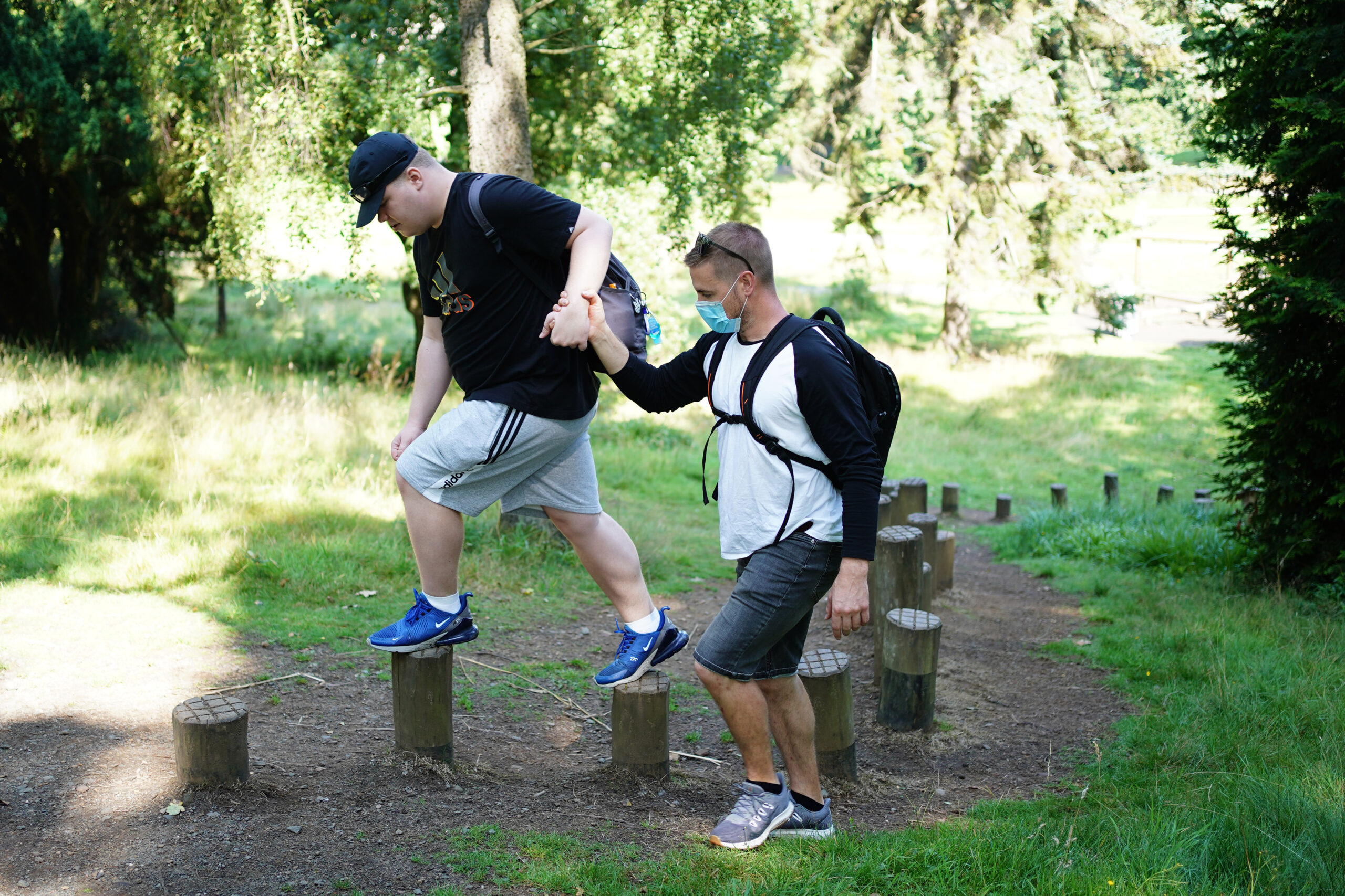 A man walks over stepping poles while holding hands for support with a man next to him. Carr Gomm