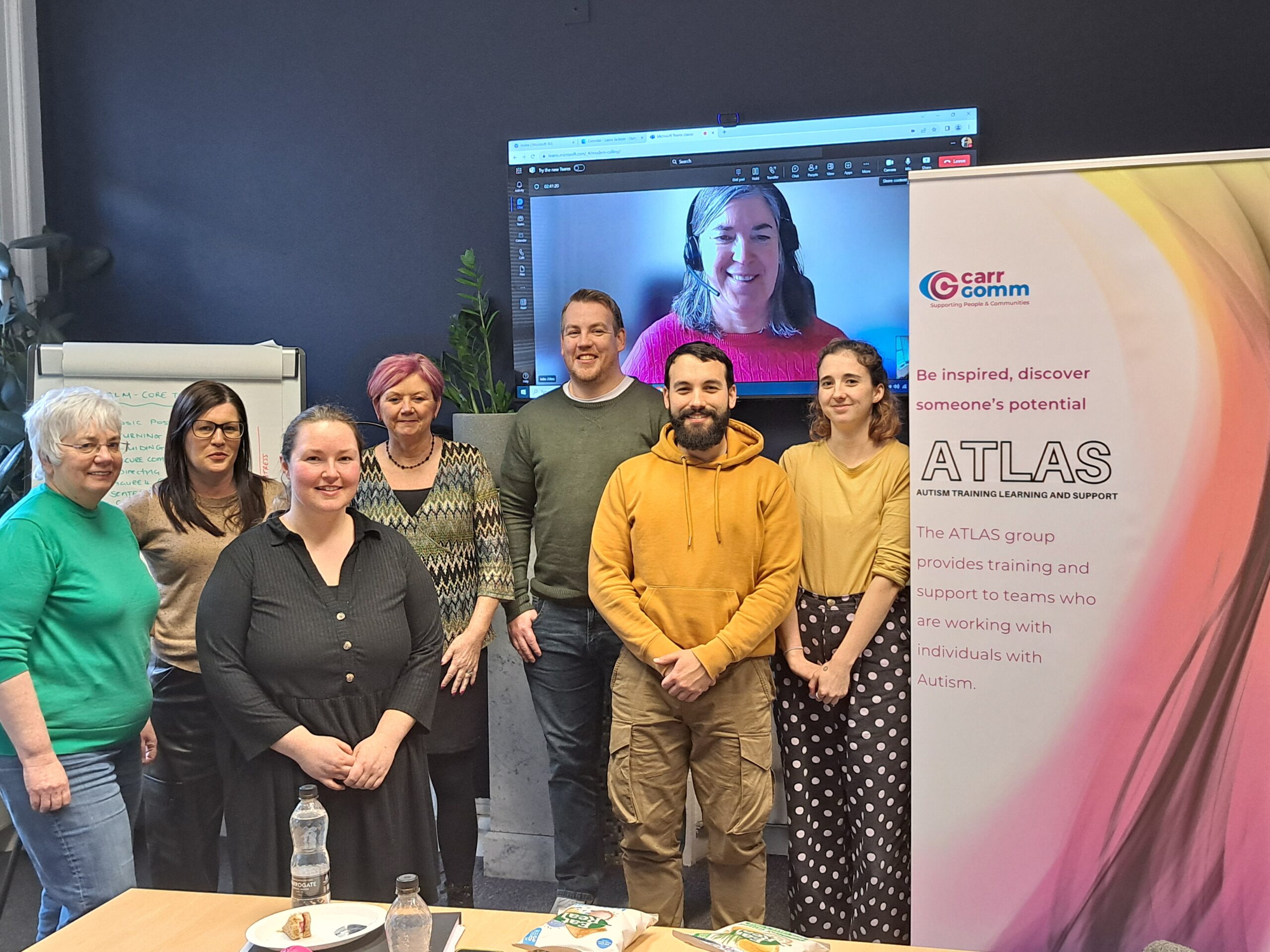 A group of people standing in front of a screen in an office environment. There is a roller banner promoting ATLAS. 