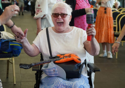An older woman (Margaret) sitting in a wheelchair laughing and waving an inflatable microphone. Carr Gomm