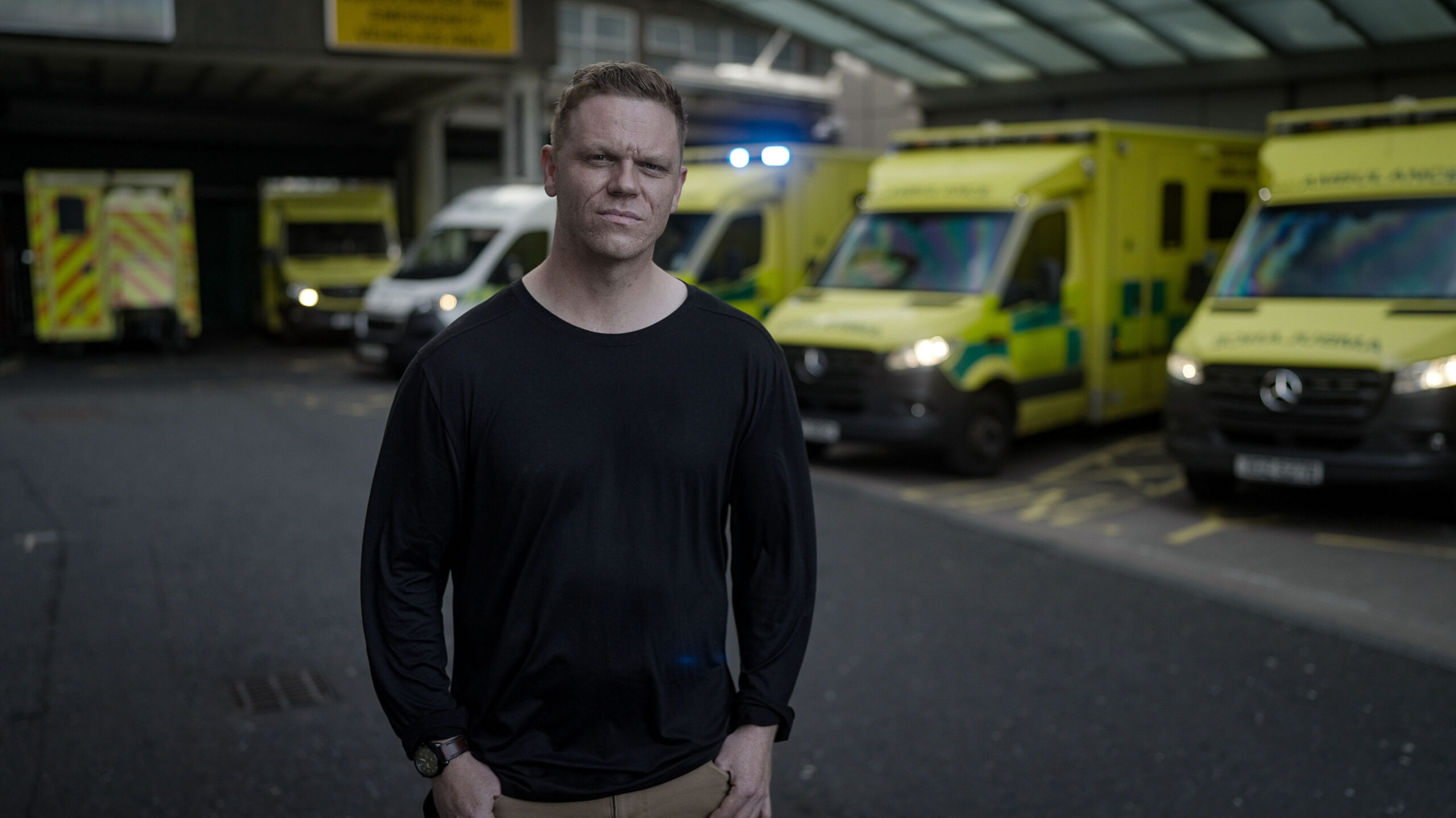 Man dressed in dark clothing stood in front or row of Ambulances outside of a hospital