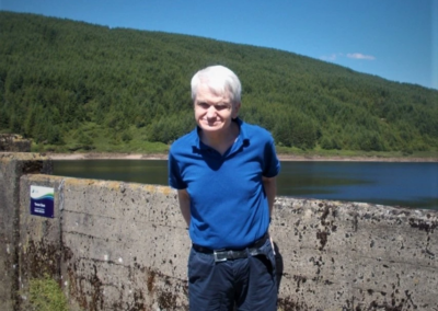 A man (James) stands for a picture in front of a resevoir. There are blue skies and dense forest in the background.
