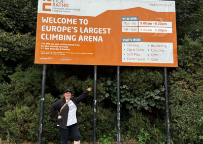 A woman (Cara) poses under an orange sign which reads 'Welcome to Europe's Largest Climbing Arena'