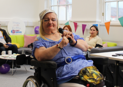 A woman in a blue shirt (Linda) in a wheelchair is dressed up like a member of ABBA and is performing a song