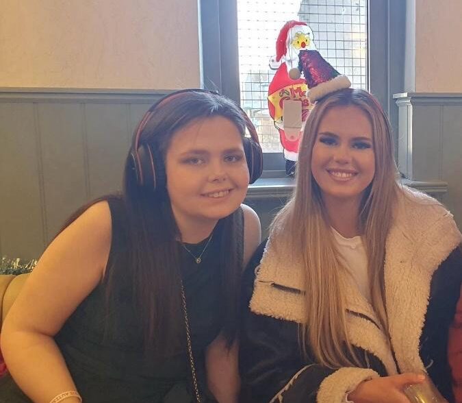 Two white women are sitting smiling at the camera. One is wear a santa hat head band and the other is wear headphones.