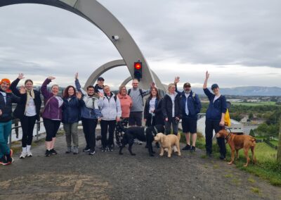A group photo of thirteen people with their arms raised in front of the Falkirk Wheel. There are also three dogs!