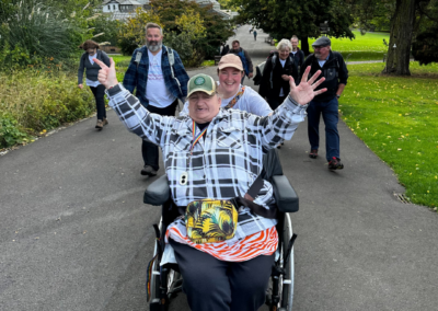 A group photo of people walking up a hill in the Glasgow Botanic Gardens. At the front of the group is a woman in a wheelchair (Linda) cheering, with her arms raised. Another woman (Cara) is beaming behind her.