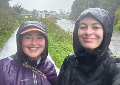 Two women (Cara and Kat) are wearing raincoats and smiling at the camera, it is raining hard!