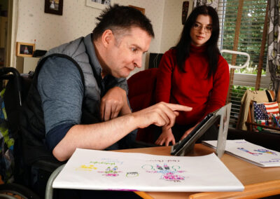 A man in a wheelchair and a woman sitting at a table with a laptop. Carr Gomm