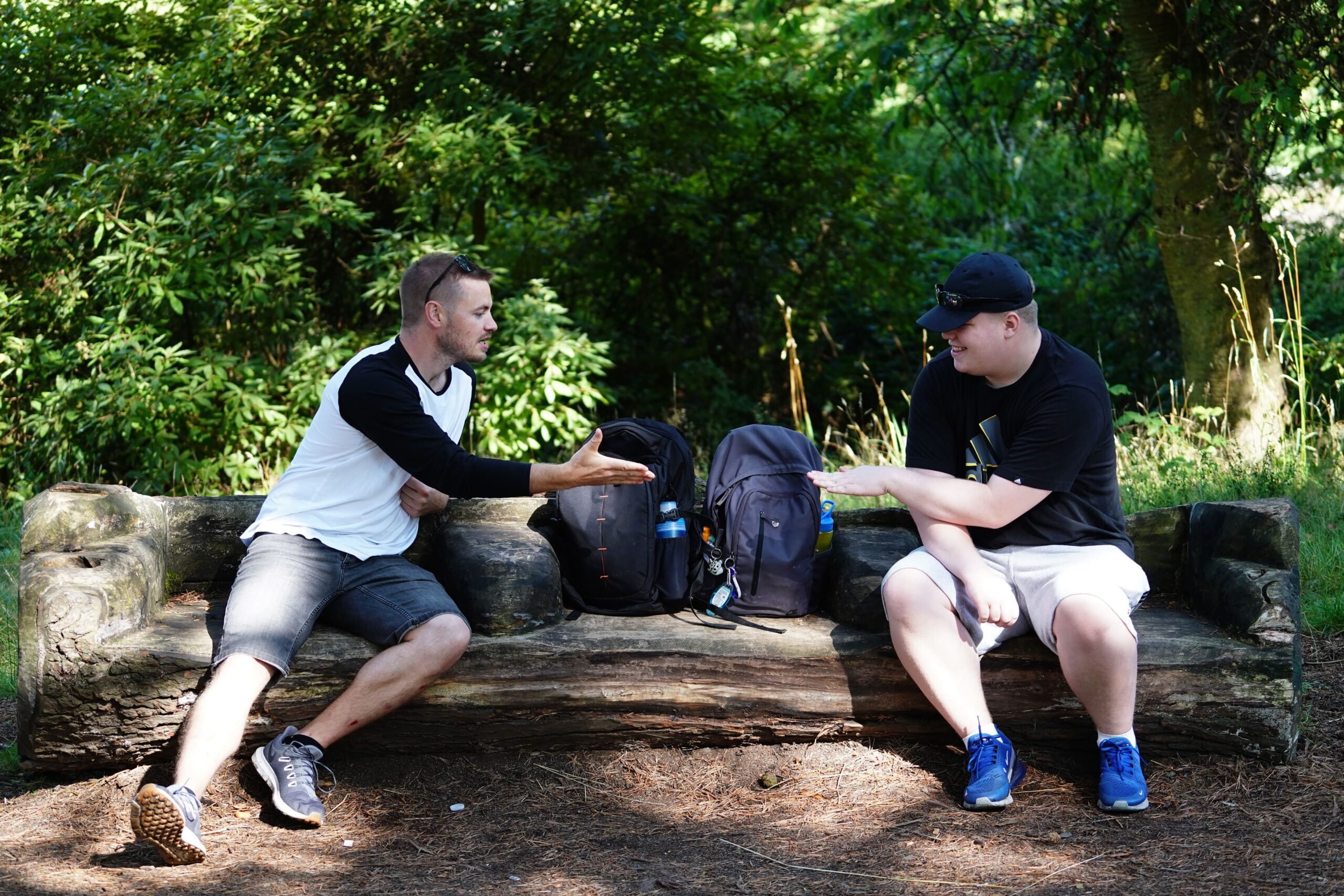 Two men sit on a log in the wooden log in a forest, and are reaching their hands out to each other