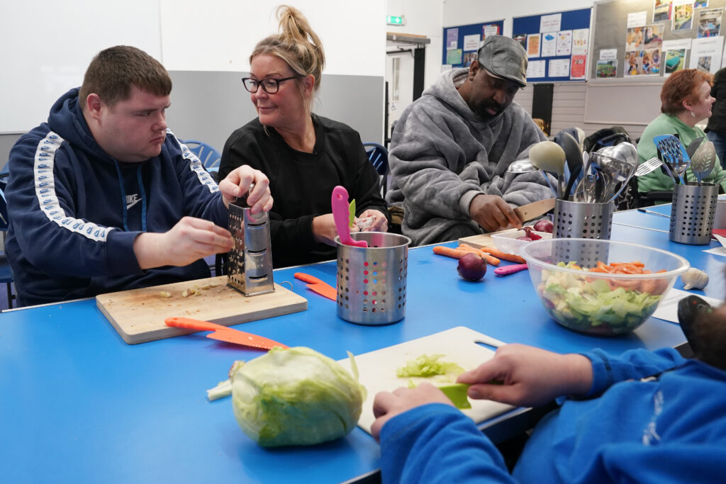A group of people participating in a cooking class, chopping vegetables and grating cheese at a table. Carr Gomm