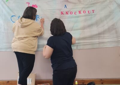 A women is painting on a sheet hung on a wall and a women is kneeling next to her helping.