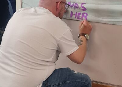 A man kneels on the floor painting onto a sheet hung on the wall. He is painting the words 'Carr Gomm was here'