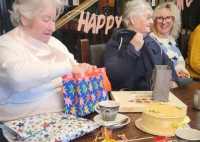 A woman (Maureen) opening a gift at a table decorated with a 'Happy Retirement' sign, with a coffee cup and other wrapped gifts nearby | Maureen’s retirement celebration after over two decades of working in the social care sector.