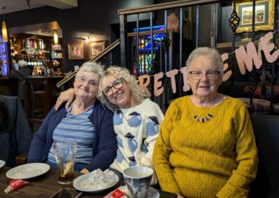 Three people smiling at a table in a café with cups visible, warm lighting, and a "Retirement" banner | Maureen’s retirement celebration after over two decades of working in the social care sector.