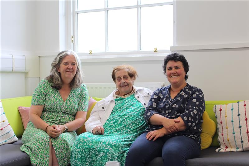 Three women sit under a sunny window smiling at the camera. NCS.
