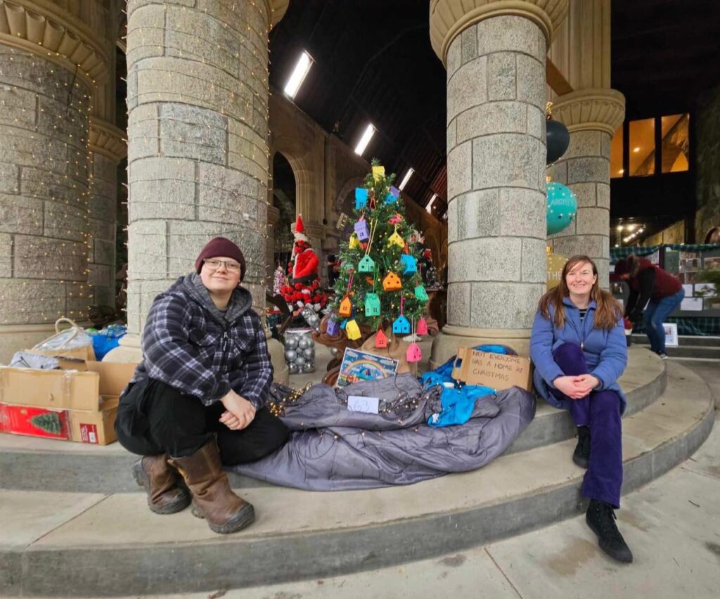 Two women sitting by a decorated Christmas tree in a stone archway, surrounded by gift boxes and holiday decorations.