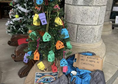 A Christmas tree decorated with colorful house-shaped ornaments and tags, positioned beside a column. A sign reading "Not everyone has a home at Christmas" is visible.