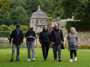 Five people walk while talking to each other. On a grassy lawn.
