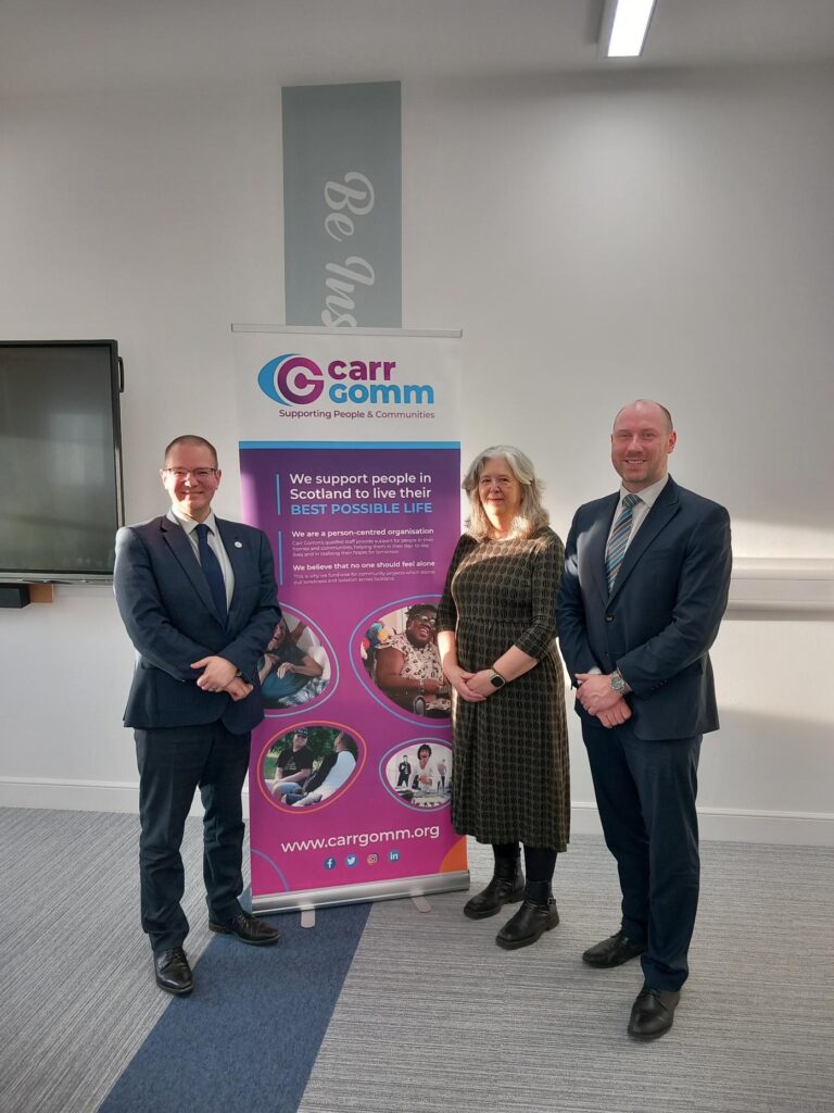 Two men (Neil Gray - Cabinet Secretary for Health and Social Care and Andrew Thomson - Deputy Chief Executive of Carr Gomm) and a women (Lucy Wren - Chief Executive of Carr Gomm) stand in front of a Carr Gomm banner.
