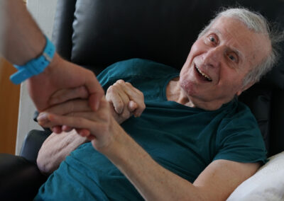 An older man is sitting in a chair smiling and holding the hand of someone out of frame.