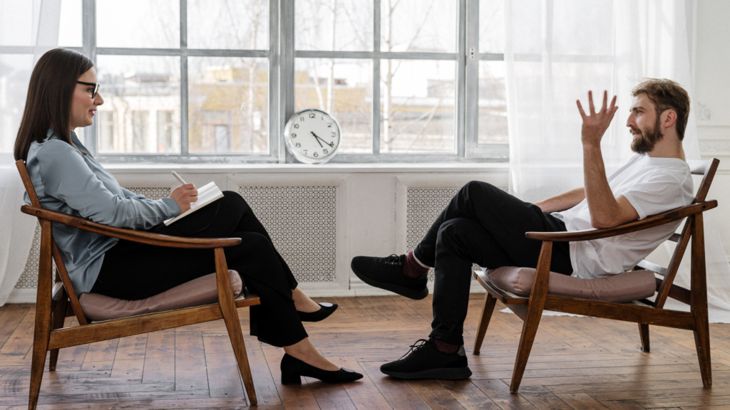 Two individuals are engaged in a conversation in a bright room with large windows. One is seated with a notepad, listening, while the other gestures expressively.