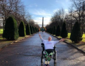 A women in a wheelchair smiles and waves her arms at the camera in Glasgow Green.