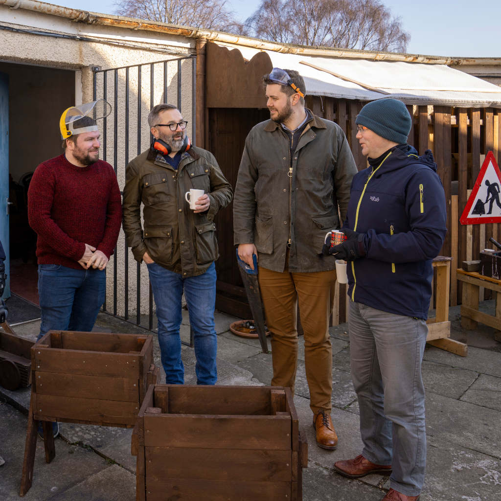 Four men stand outside a workshop chatting and smiling. | Carr Gomm