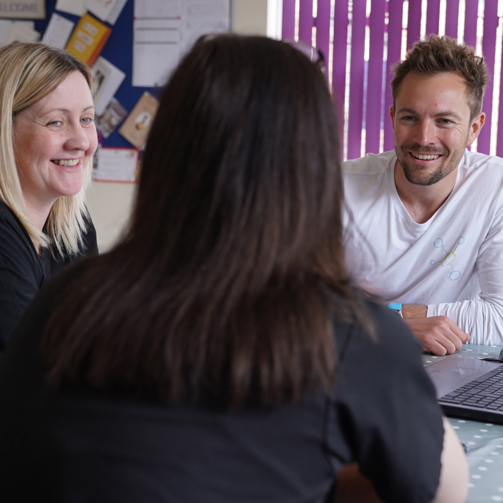 Three people seated around a table chatting. A smiling person has their back to the camera, while Fiona, Operations Manager, and Stuart, a Director on the Board, both look toward the camera