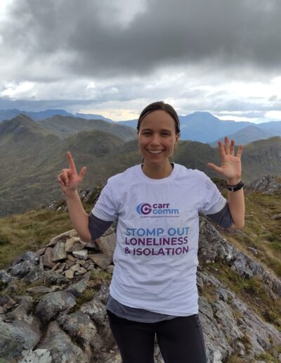Person wearing a "Carr Gomm - Stomp Out Loneliness & Isolation" t-shirt, standing on a mountain with storm clouds overhead, smiling and making a peace sign with both hands