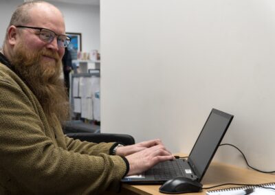 Individual with a beard smiling while working on a laptop at a desk in an office setting.