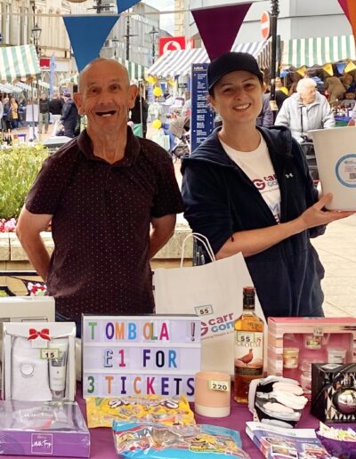 Two people standing behind a stall at a charity event, smiling and surrounded by prizes and signs promoting a tombola with tickets priced at £1 for 3. There are various items for sale displayed on the table. The setting includes outdoor decorations and a bustling, festive atmosphere.