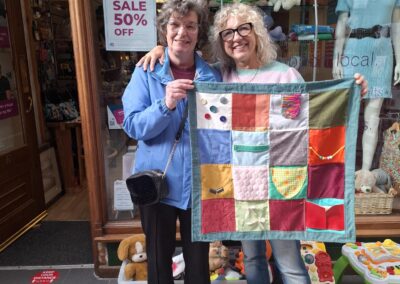 Two people standing behind a stall at a charity event, smiling and surrounded by prizes and signs promoting a tombola with tickets priced at £1 for 3. There are various items for sale displayed on the table. The setting includes outdoor decorations and a bustling, festive atmosphere.