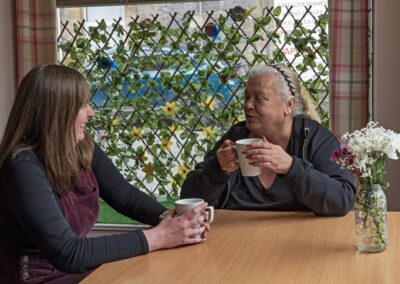 Two people enjoying a conversation over cups of tea at a table, with a window featuring a decorative lattice and plants in the background.