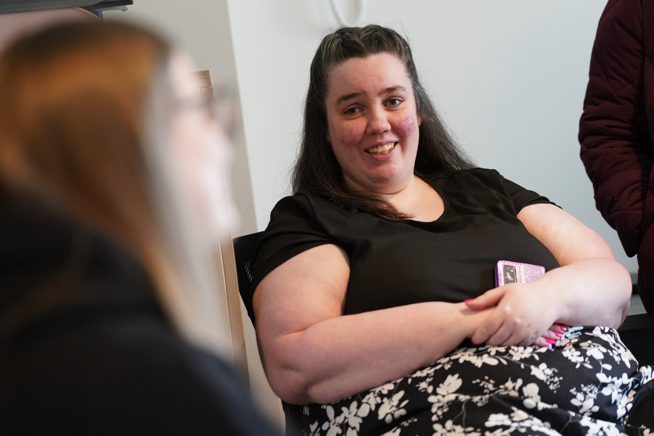 Person smiling and having a conversation in a room with another individual partially visible in the foreground.