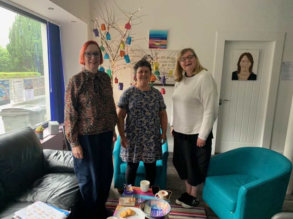 Three woman (Eilidh, Becs and Jenni) standing in a room with a small table displaying refreshments. Behind them is a decorative tree with hanging ornaments and a colorful artwork on the wall.
