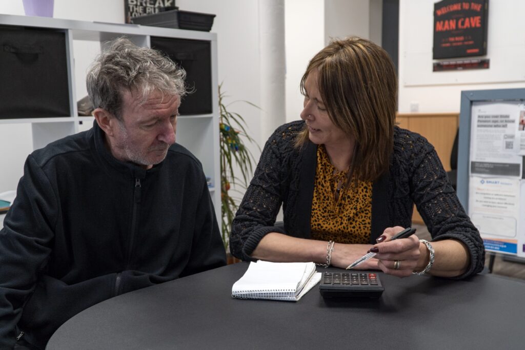 Generate by AI Assist Two individuals engaged in a discussion at a table with notepads and a pen, in a room with various posters and a plant in the background.