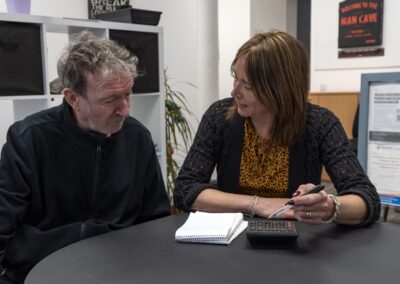 Generate by AI Assist Two individuals engaged in a discussion at a table with notepads and a pen, in a room with various posters and a plant in the background.