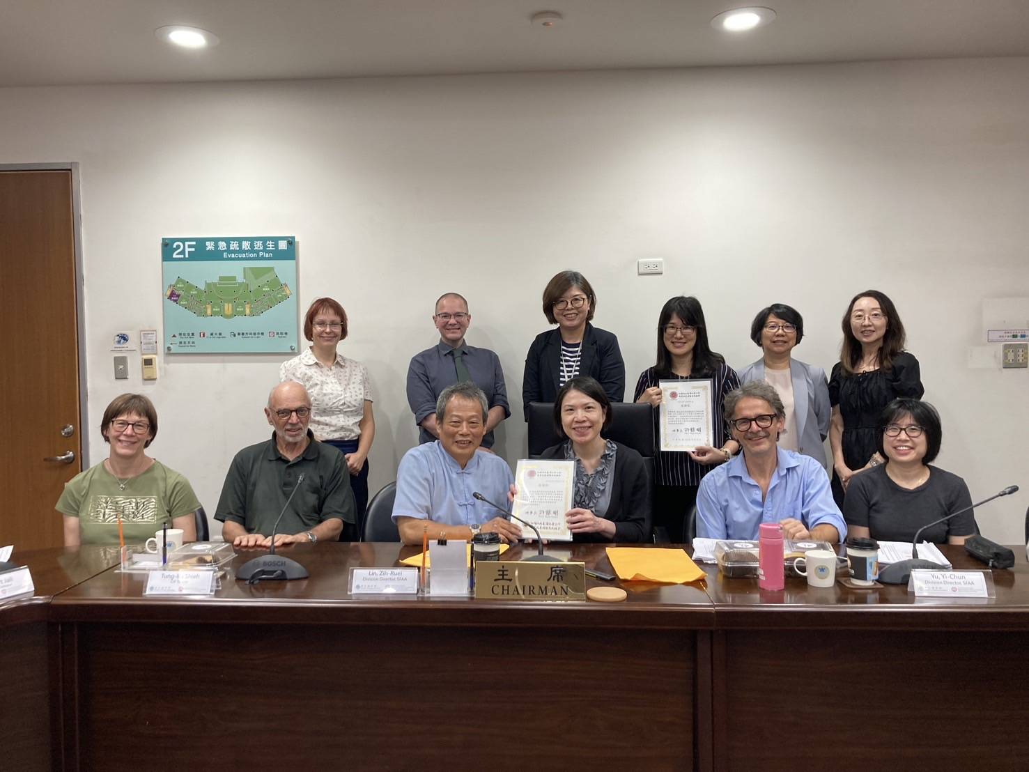 Two row of people behind a desk are smiling at the camera with some holding certificates