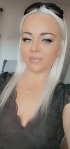 A young woman with long blonde hair wearing a black lace top and sunglasses on their head smiles for a selfie