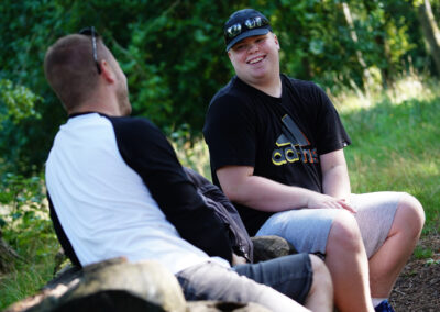 Two men sitting on a log in a park. One man faces the camera and is smiling.