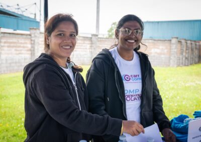 Two individuals smiling at a community event, one in a black jacket and the other wearing a white T-shirt with 'Carr Gomm' logo, standing by a table with informational material.