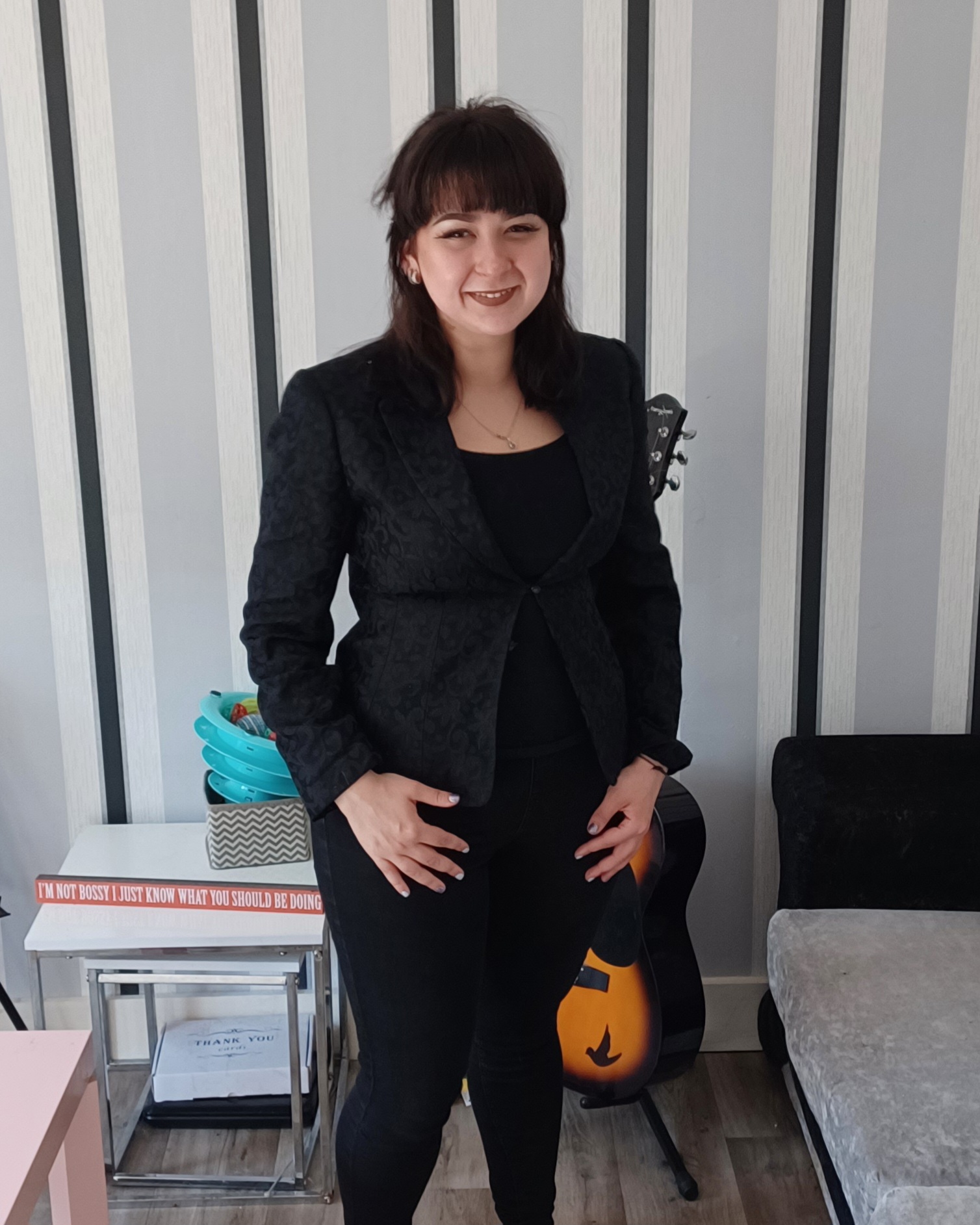 a woman (Charley) in black pants and a blazer standing next to a guitar