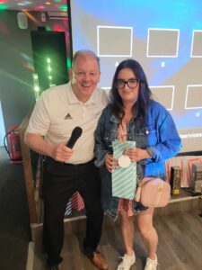 Two people smile at the camera. One is holding a microphone and the other a gift bag.