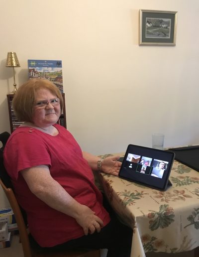 Person sitting at a table with an iPad displaying a video call screen, in a room with a framed picture and a lamp.