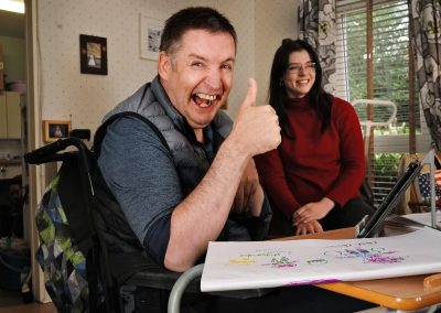 Person in a wheelchair giving a thumbs up at a table with colorful drawings, beside another person smiling, in a room with floral curtains. | Digital Inclusion Development Project